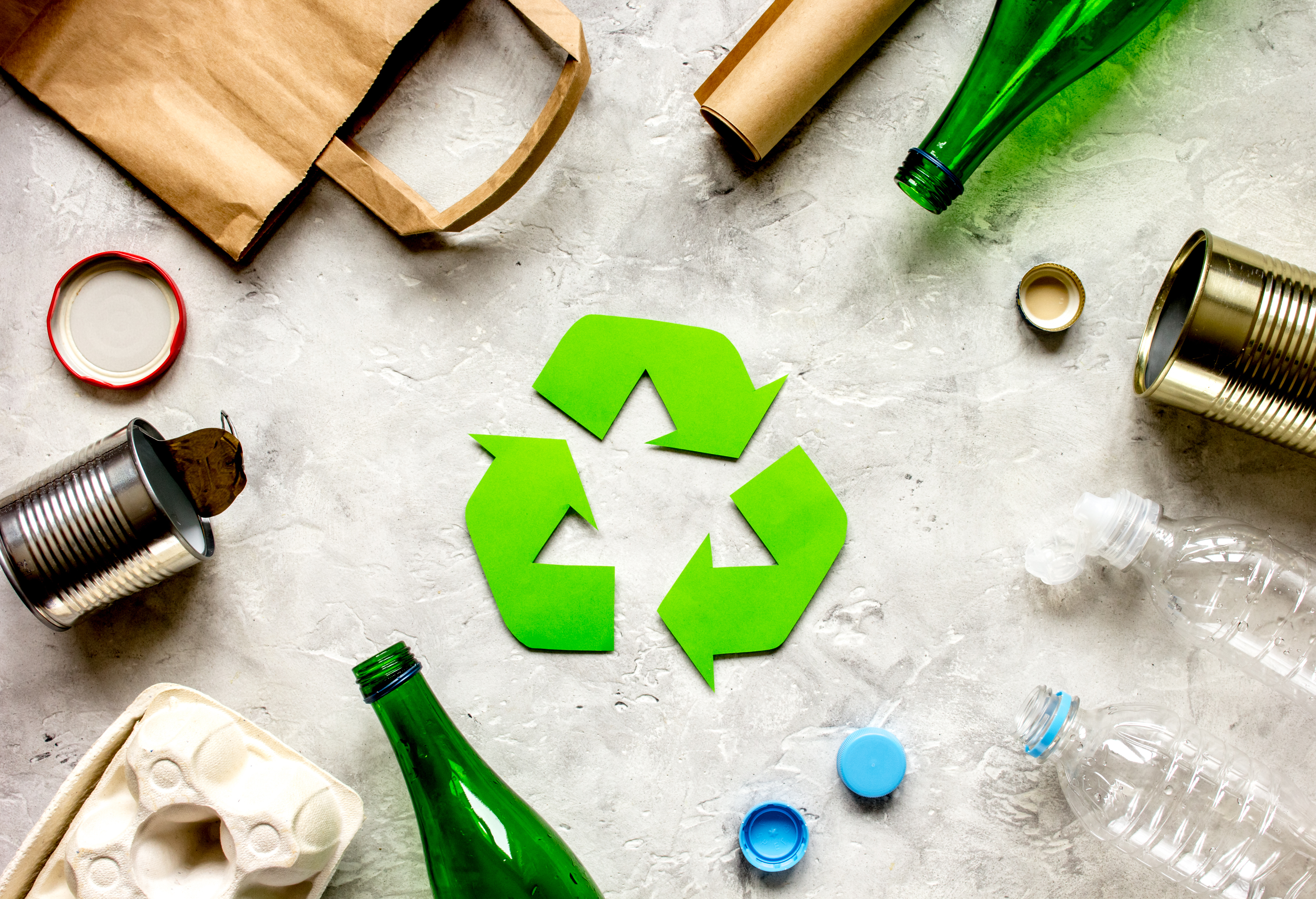Grünes Recycling-Symbol umgeben von verschiedenen Verpackungsmaterialien wie Papiertüten, Glasflaschen, Dosen und Kunststoffbehältern auf grauer Oberfläche.